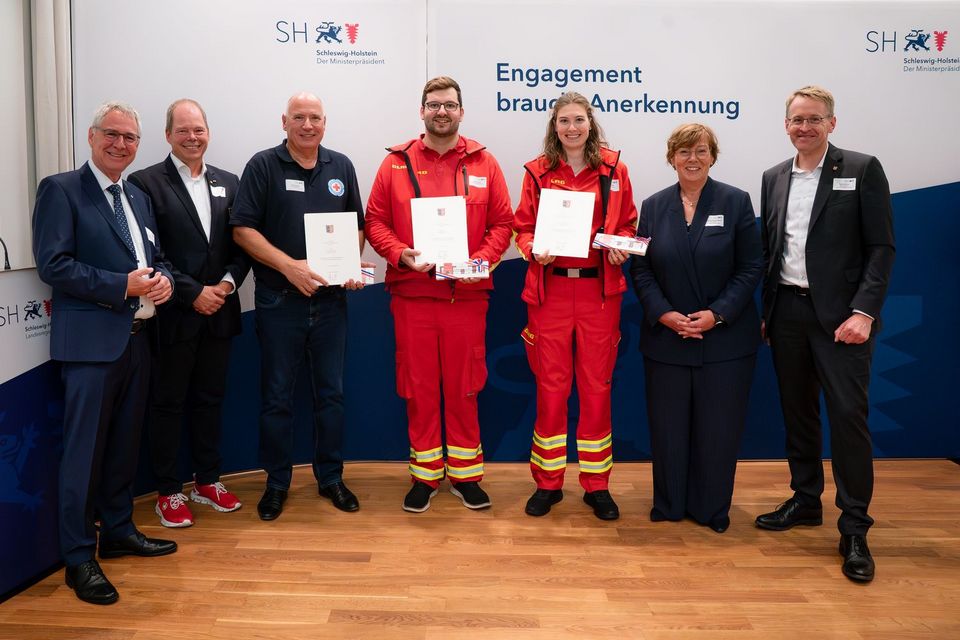 Sieben Personen stehen nebeneinander vor einer Foto-Wand, auf der steht: Engagement braucht Anerkennung.