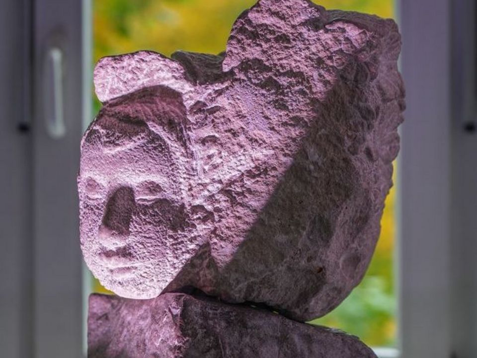 Eine steinerne helle Skulptur mit einem Gesicht steht auf einem schwarzen Sockel. Das Gesicht wird von einer Lampe angetrahlt.