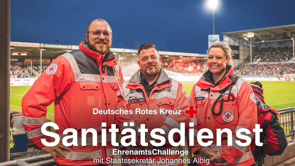 Drei Menschen in orangefarbenen DRK-Einsatzjacken stehen in einem Fußballstadion.