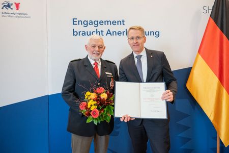 Zwei ältere Männer in Anzügen posieren mit Blumenstrauß und Urkunde für ein Foto.