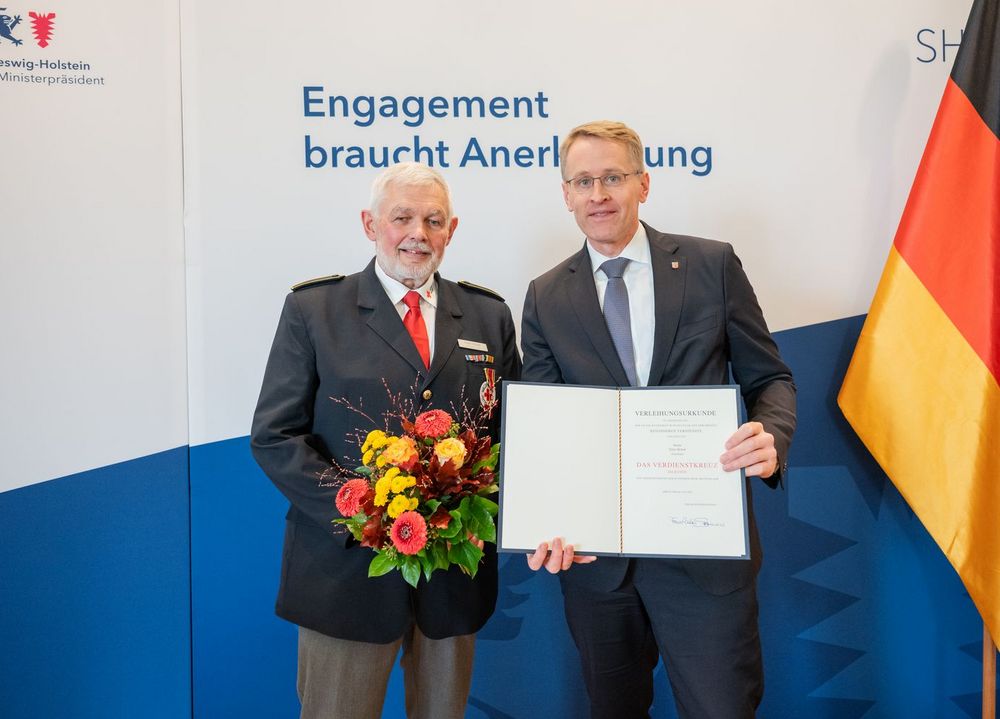 Zwei ältere Männer in Anzügen posieren mit Blumenstrauß und Urkunde für ein Foto.