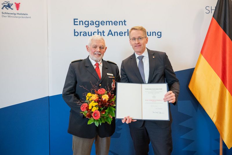 Zwei ältere Männer in Anzügen posieren mit Blumenstrauß und Urkunde für ein Foto.
