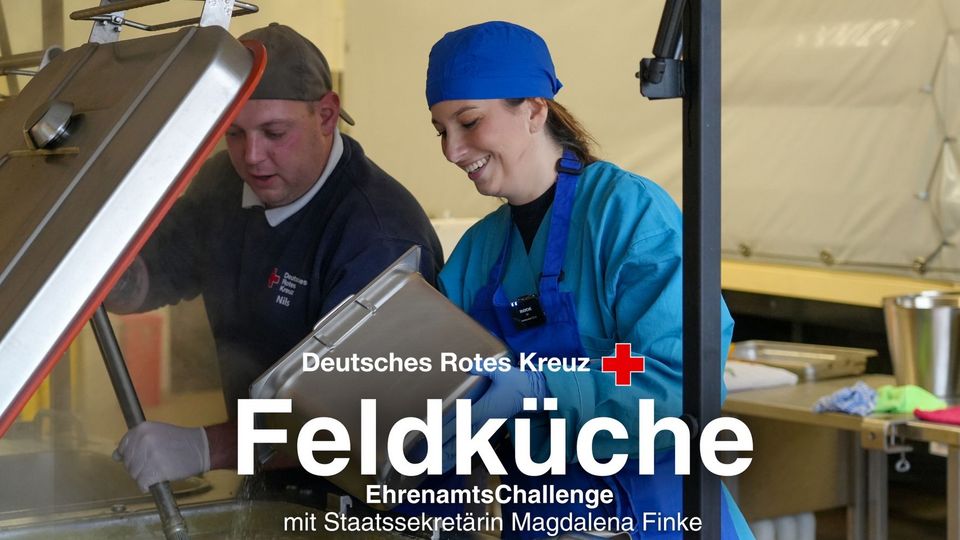 Ein junger Mann und eine junge Frau stehen nebeneinander an einem großen Topf. Darunter steht: DRK Feldküche.
