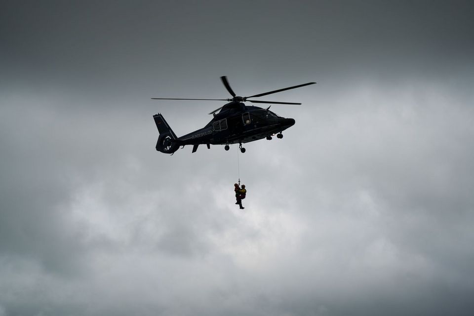 Ein Hubschrauber in der Luft: Unter ihm hängen 2 Luftretter*innen an einem Seil, an dem sie gerade wieder an Bord gezogen werden.