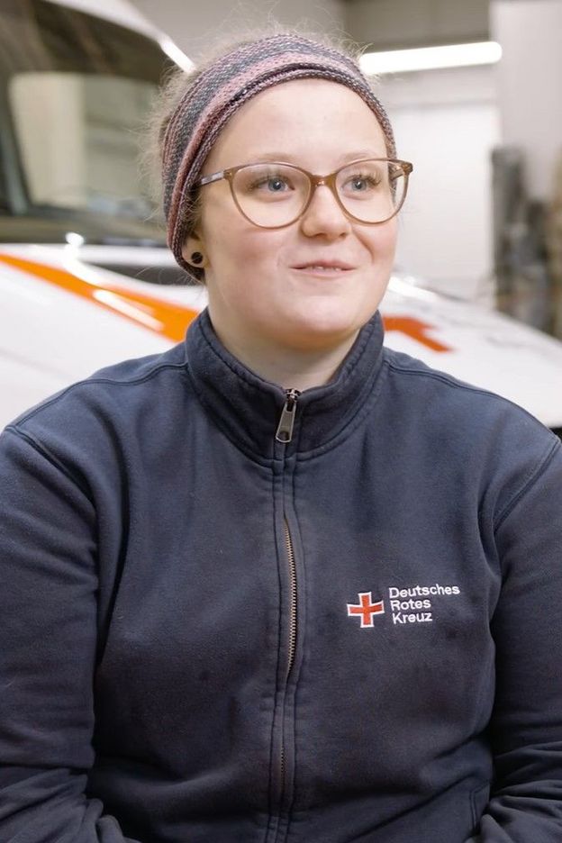 Eine junge Frau sitzt in einer Halle vor einem Fahrzeug. Sie trägt eine blaue Jacke mit der Aufschrift "Deutsches Rotes Kreuz".