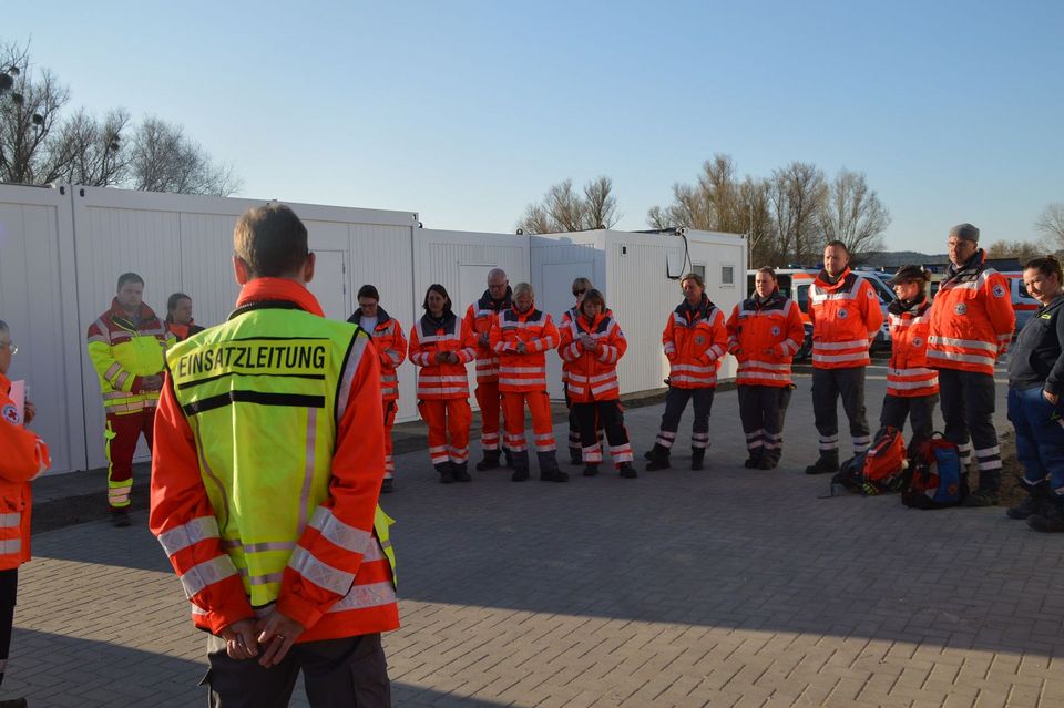 Ca. 20 Menschen in roten DRK-Einsatzjacken stehen im Halbkreis der "Einsatzleitung" gegenüber, die eine damit beschriftete gelbe Warnweste trägt.
