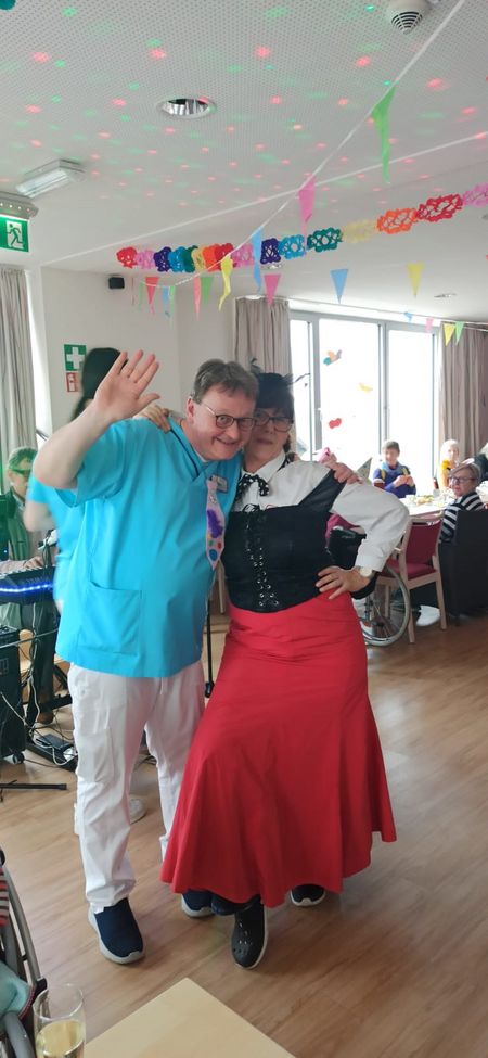 Ein Mann mit Jecken-Hut hält eine Rede in einem geschmückten Saal, in dem zahlreiche Senior*innen sitzen.
