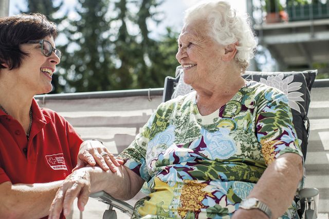 Eine lächelnde Seniorin sitzt auf einem Gartenstuhl in der Sonne. Neben ihr kniet eine Pflegekraft des DRK.