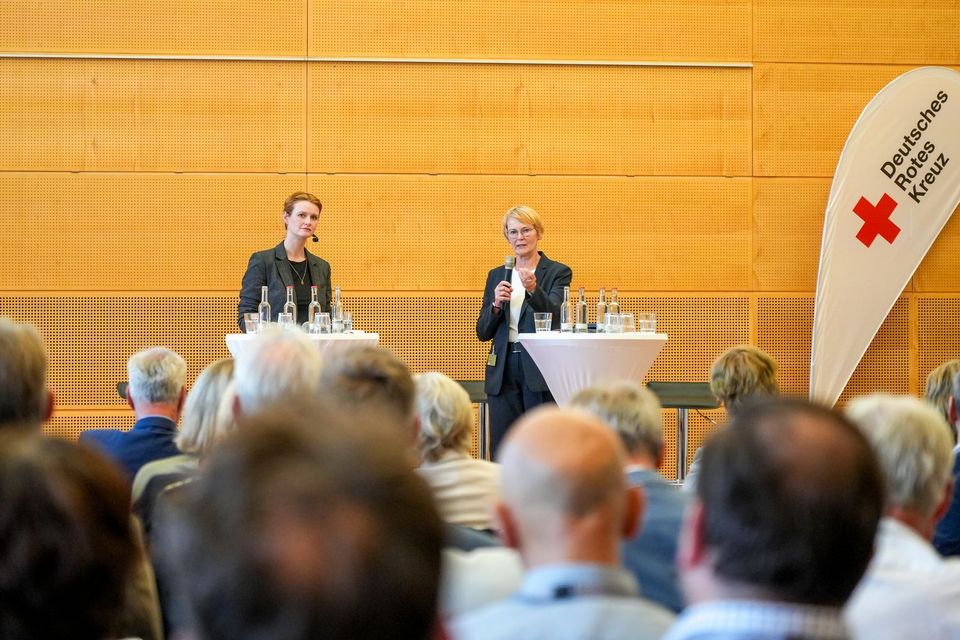 Zwei Menschen stehen auf einer Bühne und richten sich an das Publikum im Vordergrund.